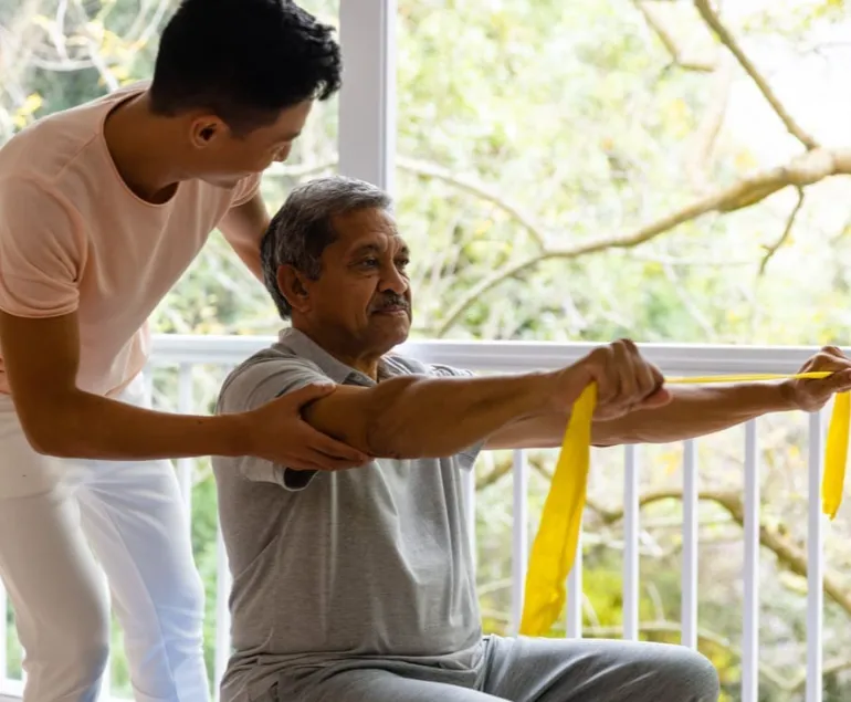 Indian-elderly-man-performing-rehabilitation-exercises-using-resistance-bands,-assisted-by-a-physiotherapist