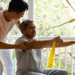 Indian-elderly-man-performing-rehabilitation-exercises-using-resistance-bands,-assisted-by-a-physiotherapist