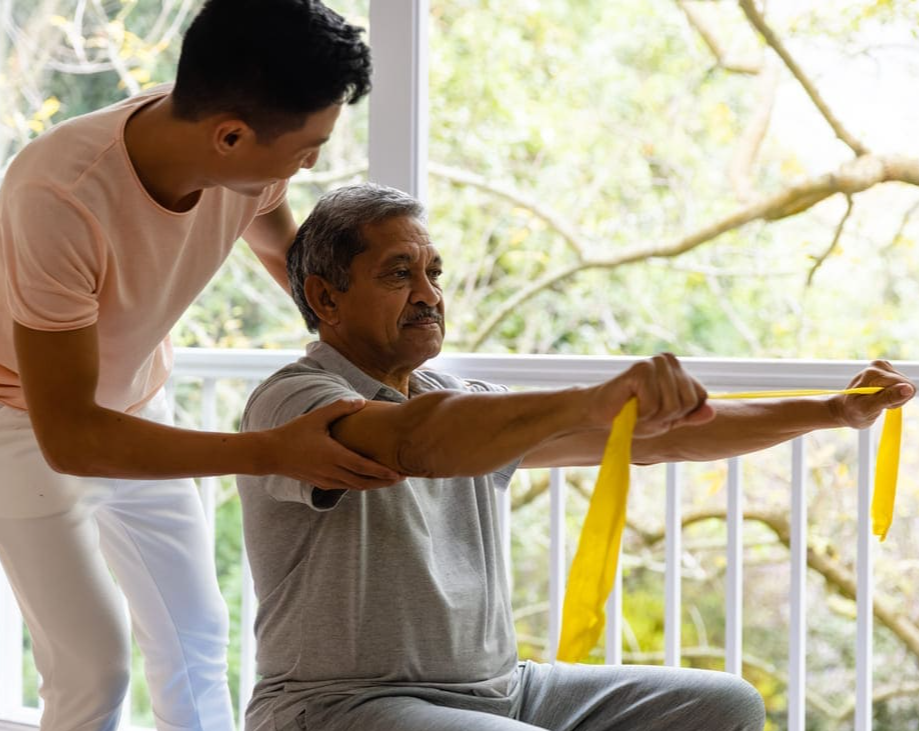 Indian-elderly-man-performing-rehabilitation-exercises-using-resistance-bands,-assisted-by-a-physiotherapist